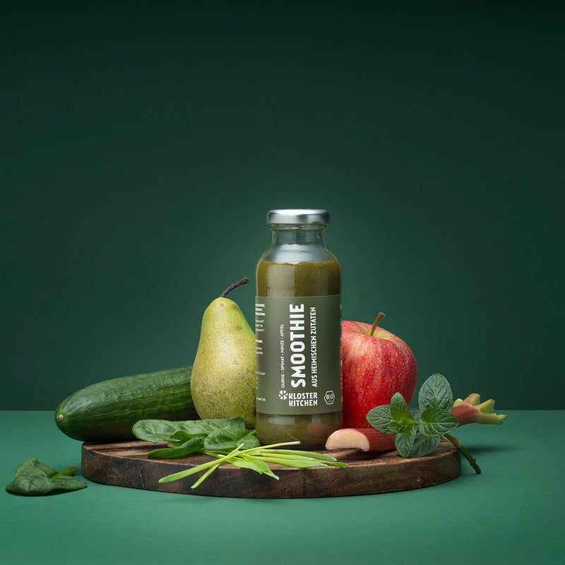 Smoothie concombre épinard menthe pomme sur un fond vert. Le smoothie est posé sur une planche en bois et les ingrédients du smoothie sont alignés à côté. Appuyés contre la bouteille se trouvent : Concombre, menthe, poire, pomme, rhubarbe.