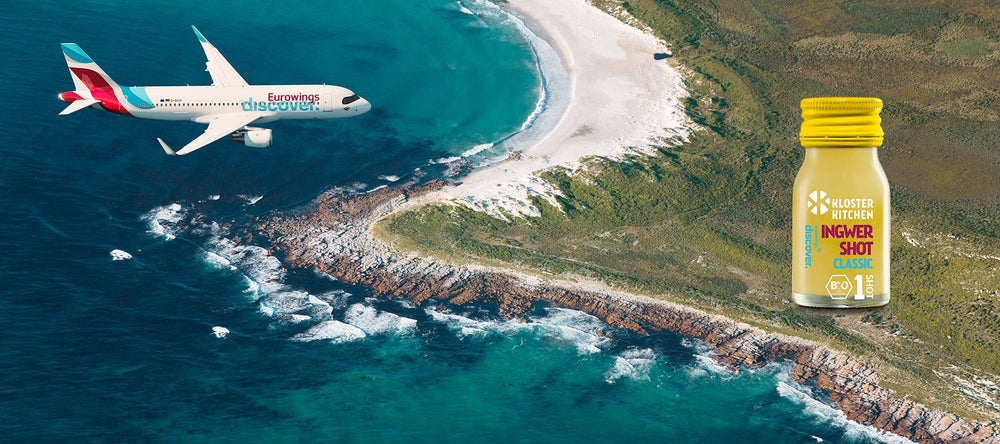 Ein Flugzeug der Eurowings Discover über einer Küste. Auf dem Festland steht ein Ingwer Shot Classic im Eurowings Discover Design.