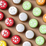 Diseño plano: tapones de botellas redondos y coloridos sobre fondo marrón, dispuestos en forma de cuadrícula con tapones parcialmente superpuestos; rojo, verde, naranja y plateado, algunos con motivos de rodajas de cítricos blancas, otros con brillo metálico.