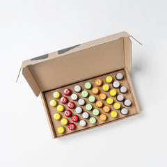 Offener brauner Karton mit 48 bunten, runden Kappen in einem 8×6-Raster; Farben wie Rot, Gelb, Grün, Orange und Grau mit sternförmigen Mustern, auf hellem Hintergrund.