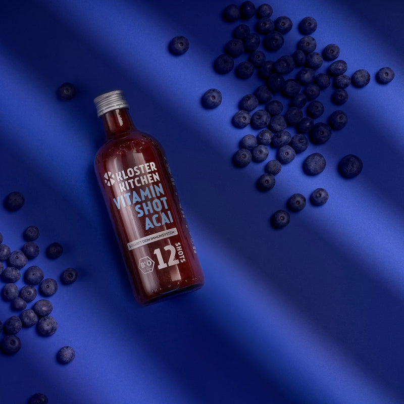 The Vitamin Shot Acai 12SHOTS from Kloster Kitchen, lying on a blue background. Around the 360 ml bottle are distributed a few handfuls of acai berries. 