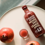 1 von den 3x 12SHOTS 360 ml Ingwer Shot Granatapfel: die Flasche steht auf einem Tisch, daneben steht ein eingeschenkter Ingwer Shot in einem Shot Glas. Daneben steht ein Granatapfel.