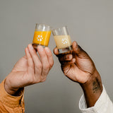 Two filled shot glasses from Kloster Kitchen are held by two hands as if they are about to toast.