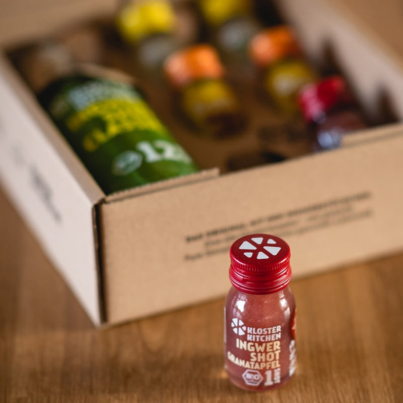 In the background, the Ginger Shot Tasting Set S can be seen blurred open on a wooden table. In the front, the focus is on a single Ginger Shot Pomegranate, which was taken from the tasting set.
